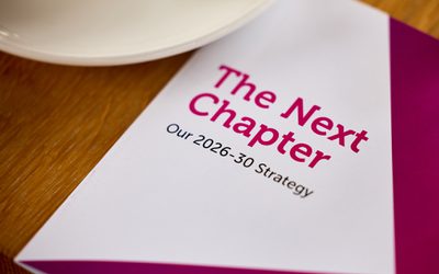 Booklet on a wooden table. The front cover is white and pink and the title reads "The Next Chapter Our 2026-30 Strategy"