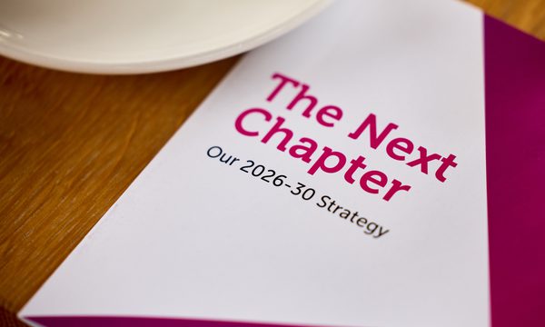 Booklet on a wooden table. The front cover is white and pink and the title reads "The Next Chapter Our 2026-30 Strategy"