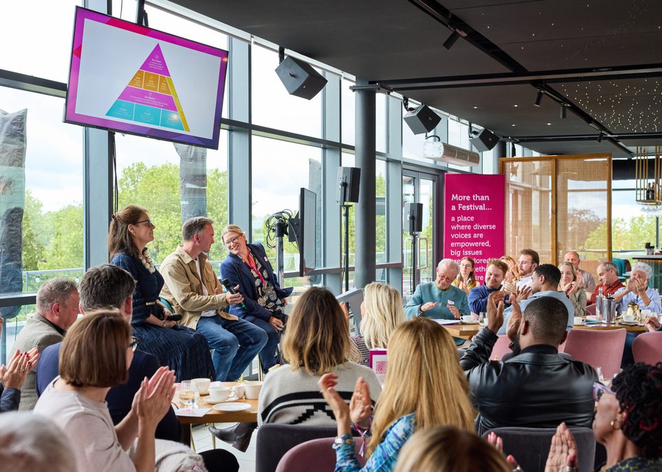 Group of people sat down clapping following a speech by 3 people who are at the front of the room. A TV screen hung high up shows a pyramid of different colours illustrating the 2026-2030 strategy