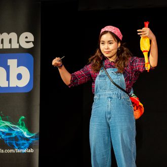 A FameLab participant stands on stage during their talk holding a rubber chicken